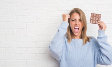 Güzel ve genç birbayan yeme çikolata bar rahatsız ve sinirli öfkeyle, çılgın ve yükseltilmiş eliyle diye bağıran öfke kavramı bağırarak beyaz tuğla duvarın üzerinden