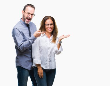 Orta yaş İspanyol çift hayrete ve el ile sunulması ve parmak ile işaret ederken kameraya gülümseyen izole arka plan üzerinde gözlük takan aşık.