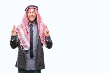 Genç yakışıklı Arap adam uzun saçlı geçiş parmakları umut ve gözleri kapalı ile gülümseyen izole arka plan üzerinde keffiyeh giyiyor. Şans ve batıl kavramı.