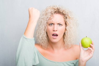 Genç Sarışın kadın yüzünden grunge gri arka plan yeşil elma rahatsız yemek ve boşa çıkarmak ile öfke, çılgın ve yükseltilmiş eliyle diye bağıran öfke kavramı bağırarak.