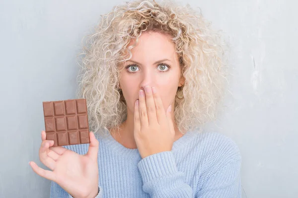 Young Blonde Woman Grunge Grey Wall Eating Chocolate Bar Cover — Stock Photo, Image