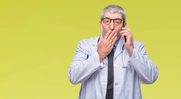 Handsome senior doctor man talking on smarpthone over isolated background cover mouth with hand shocked with shame for mistake, expression of fear, scared in silence, secret concept
