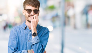 Genç yakışıklı adam stresli ve gergin ile izole arka plan üzerinde güneş gözlüğü takıyor tırnak ısırma dudaklarından eller. Anksiyete sorunu.