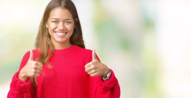 El ile pozitif jest yapıyor izole arka plan başarı işareti üzerinde kırmızı kış kazak giyen genç güzel esmer kadın başparmak gülümseyen ve mutlu. Neşeli bir ifadeyle, kazanan jest kameraya bakıyor.