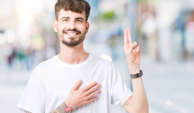 Beyaz t-shirt üzerinde izole giyen genç yakışıklı adam arka plan Swearing göğüs ve parmaklar, el ile bir sadakat sözü yemin yapma