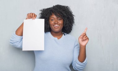 Gri grunge genç Afro-Amerikan kadın boş kağıt sayfası sürpriz bir fikir ile tutarak duvar veya gülen yüz, bir numara ile işaret parmağı soru