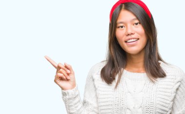 Kameraya bakarak yan el ve parmak ile işaret yüzünde kocaman bir gülümseme ile izole arka plan üzerinde kış kazak giyen genç Asyalı kadın.