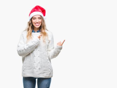 Gülümseyen ve iki elleri ve parmakları ile tarafına işaret kamera bakarak izole arka plan üzerinde Noel şapka giyen güzel genç sarışın kadın.