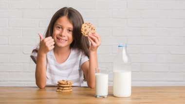 Genç İspanyol süt içme masada oturan çocuk ve çikolata bisküvi mutlu ok işareti yapıyor büyük gülümseme ile yemek kadar parmakları ile mükemmel işaret parmak
