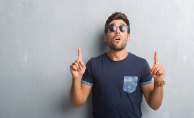 Yakışıklı genç güneş gözlüğü takıyor gri grunge duvarın üzerinden hayran ve yukarıya bakıyor ve parmakları ile işaret ve silah kaldırdı şaşırttı.