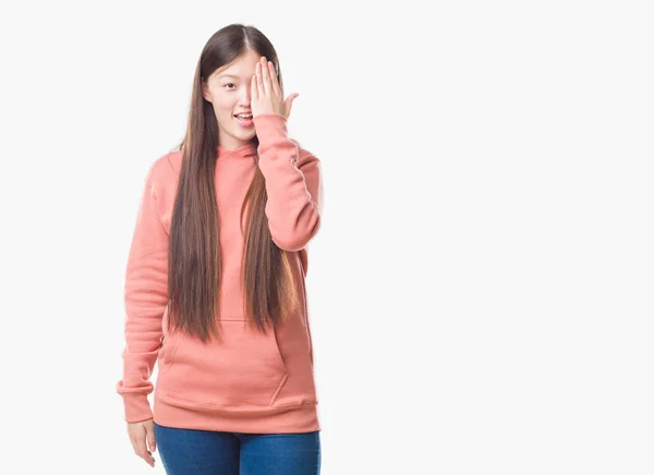 若い中国の女性を分離顔と驚きの感情に自信を持って笑顔で手で片目を覆うスポーツ Sweathshirt を身に着けている背景 — ストック写真