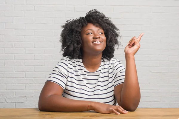 Young african american woman sitting on the table at home with a big smile on face, pointing with hand and finger to the side looking at the camera.