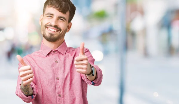 若いハンサムな男の手で肯定的なジェスチャーをして承認する分離の背景にピンクのシャツを着て笑顔と幸せな成功のための親指します カメラ 勝者のジェスチャーを見てください — ストック写真
