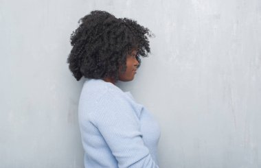 Gri grunge duvar tarafına, seyir kış kazak giyen genç Afro-Amerikan kadın dinlenmek doğal yüz emin gülümseme ile pozuyla profili.