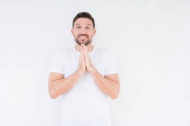 Ellerle birlikte AF emin gülümseyen için soran dua izole arka plan üzerinde rahat beyaz t-shirt giyen genç yakışıklı adam.