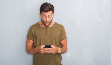 Yakışıklı genç gri grunge üzerinde duvar manifatura bir sürpriz yüz, korkuyor ve heyecanlı korku ifadesi ile smartphone şokta korktuğunu kullanarak bir ileti