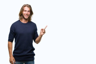 Kameraya bakarak yan el ve parmak ile işaret eden genç yakışıklı adam uzun saçlı yüzünde kocaman bir gülümseme ile izole arka plan üzerinde kış kazak giyiyor.