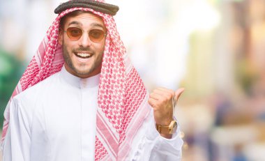 Keffiyeh seyir ve başparmak yanında yukarıyı mutlu yüz ile gülümseyen izole arka plan üzerinde giyen genç yakışıklı adam.