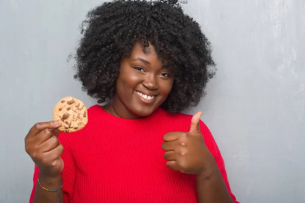 Young african american woman over grey grunge wall eating chocolate chip cooky happy with big smile doing ok sign, thumb up with fingers, excellent sign