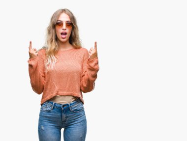 Güzel genç sarışın kadın hayrete ve yukarıya bakıyor ve parmakları ile işaret ve silah kaldırdı sürpriz izole arka plan üzerinde güneş gözlüğü takıyor.