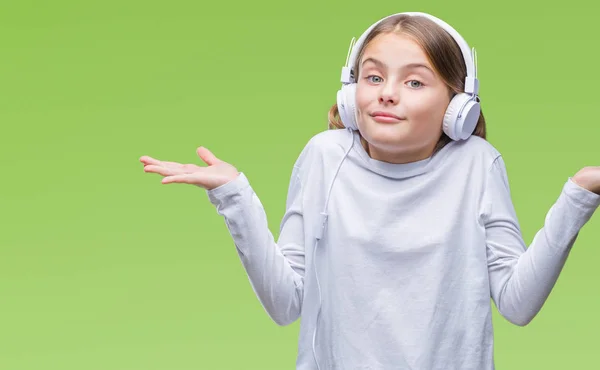 年轻美丽的女孩戴着耳机听音乐在孤立的背景无线索和混乱的表情与手臂和手抬起 怀疑概念 — 图库照片