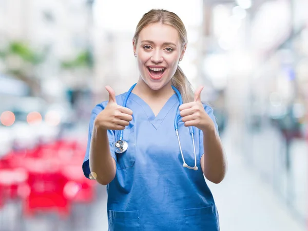 Young blonde surgeon doctor woman over isolated background approving doing positive gesture with hand, thumbs up smiling and happy for success. Looking at the camera, winner gesture.