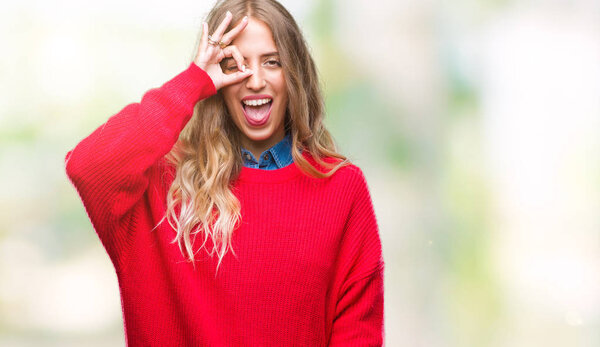 Beautiful young blonde woman wearing winter sweater over isolated background doing ok gesture with hand smiling, eye looking through fingers with happy face.