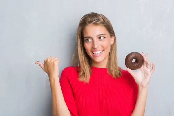 指していると幸せそうな顔を笑顔で側まで親指で示すチョコレート ドーナツを食べてグランジ灰色の壁に美しい若い女性 — ストック写真