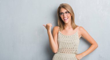 İşaret ve başparmak kadar mutlu yüz gülümseyen tarafı ile gösteren gözlük takan grunge gri duvar üzerinde duran güzel genç kadın