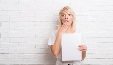 Hata, korku, sessizlik içinde gizli kavramı korkmuş ifade için utanç ile boş kağıt sayfası kapak ağız eliyle tutarak beyaz tuğla duvarın üzerinden yetişkin kafkas kadın şok