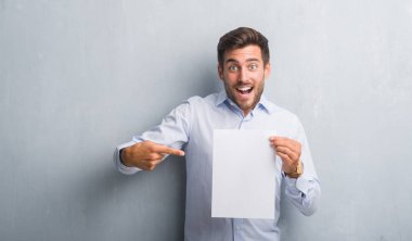 Yakışıklı genç gri grunge duvar holding boş kağıt sayfası üzerinden sözleşme çok mutlu el ve parmak ile işaret