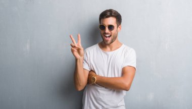 Mutlu yüz zafer işareti yaparken kameraya göz kırpıyor ile gülümseyen güneş gözlüğü takıyor gri grunge duvarın üzerinden yakışıklı genç adam. İki numaralı.