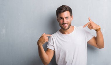 Yüzünde gülümseme ile kendinden emin görünüyorsun, kendini gururlu ve mutlu parmakları ile işaret gri grunge duvarın üzerinden yakışıklı genç.