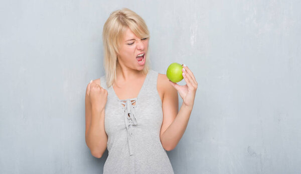 Adult caucasian woman over grunge grey wall eating green apple annoyed and frustrated shouting with anger, crazy and yelling with raised hand, anger concept