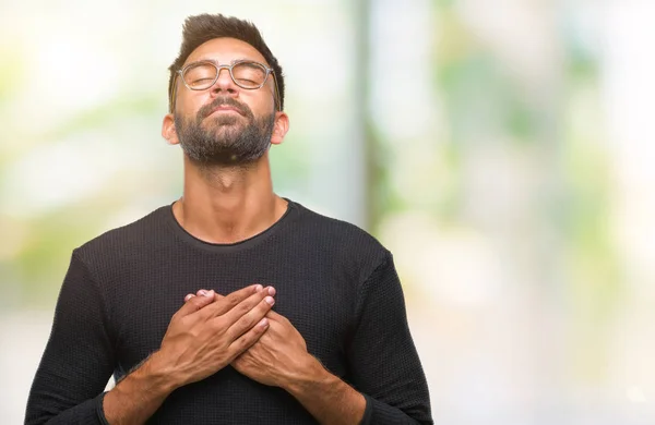 Adult hispanic man wearing glasses over isolated background smiling with hands on chest with closed eyes and grateful gesture on face. Health concept.