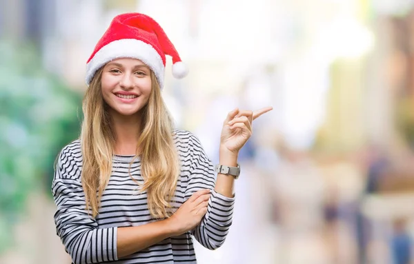 若い金髪美人の顔に大きな笑みを浮かべて隔離された背景にクリスマスの帽子を着てカメラを見て側の手と指で指す — ストック写真