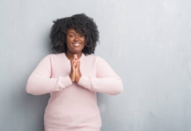 Young african american plus size woman over grey grunge wall wearing winter sweater praying with hands together asking for forgiveness smiling confident. clipart