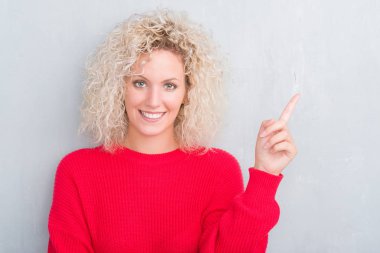 Kameraya bakarak yan el ve parmak ile işaret eden sarışın genç kadın yüzünde kocaman bir gülümseme ile grunge gri arka plan üzerinde Kıvırcık saçlı.