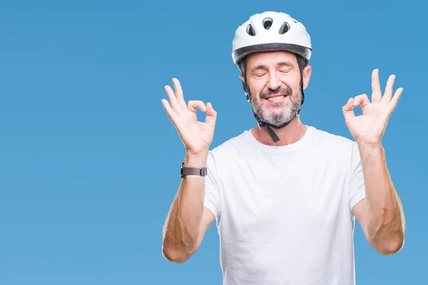 Middle age senior hoary cyclist man wearing bike safety helment isolated background relax and smiling with eyes closed doing meditation gesture with fingers. Yoga concept.