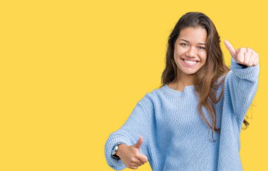 El ile pozitif jest yapıyor onaylama izole arka plan üzerinde mavi kış kazak giyen genç güzel esmer kadın başparmak gülümseyen ve başarı için mutlu. Kamera, kazanan jest arıyorsunuz.