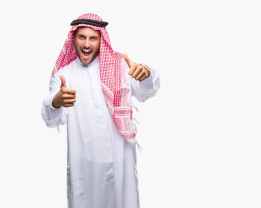 Keffiyeh el ile pozitif jest yapıyor onaylama izole arka plan üzerinde giyen genç yakışıklı adam başparmak gülümseyen ve başarı için mutlu. Kamera, kazanan jest arıyorsunuz.