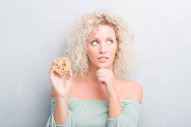 Sarışın genç kadın çikolatalı bisküvi ciddi bir yüz düşünme hakkında soru, çok karışık fikir yeme grunge gri duvarın üzerinden
