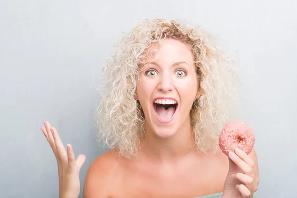 Young Blonde Woman Grunge Grey Background Eating Pink Donut Very — Stock Photo, Image