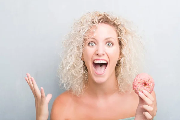 Joven Rubia Sobre Fondo Gris Grunge Comiendo Rosquilla Muy Feliz — Foto de Stock