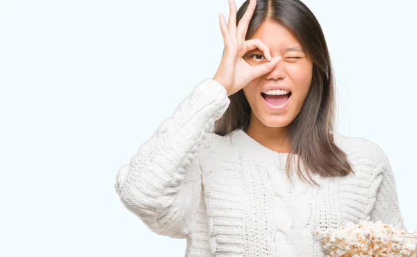 年轻的亚洲妇女吃爆米花在孤立的背景与快乐的脸微笑做 标志与手在眼睛看通过手指 — 图库照片