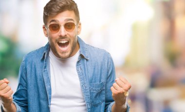 Yakışıklı delikanlı izole arka plan kutlama üzerinde güneş gözlüğü takıyor şaşırttı ve başarı için kaldırdı kollarını açarak hayret ve gözleri açık. Kazanan kavramı.