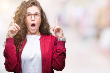 Ceket ve gözlük izole arka plan üzerinde giyen güzel esmer kıvırcık saçlı Genç kız hayran ve yukarıya bakıp parmakları ile işaret şaşırttı ve silah kaldırdı.