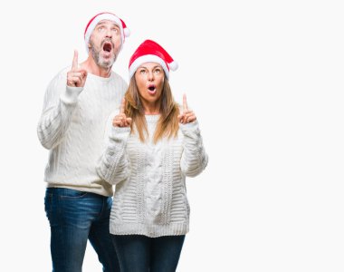 Orta yaş İspanyol çift izole arka plan üzerinde Noel şapka hayret ve yukarıya bakıp parmakları ile işaret şaşırttı ve silah kaldırdı.