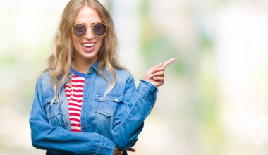 Kameraya bakarak yan el ve parmak ile işaret yüzünde kocaman bir gülümseme ile izole arka plan üzerinde güneş gözlüğü takıyor güzel sarışın genç kadın.