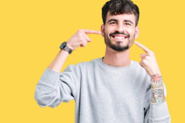 Genç yakışıklı adam kendinden emin gösterilen ve işaret parmakları diş ve ağız ile gülümseyen izole arka plan üzerinde kazak giyiyor. Sağlık kavramı.